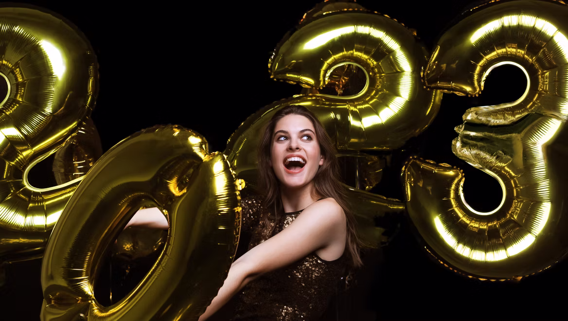 A smiling woman in a sparkly dress poses with large gold balloons shaped as the numbers "2023" against a black background, celebrating the New Year.