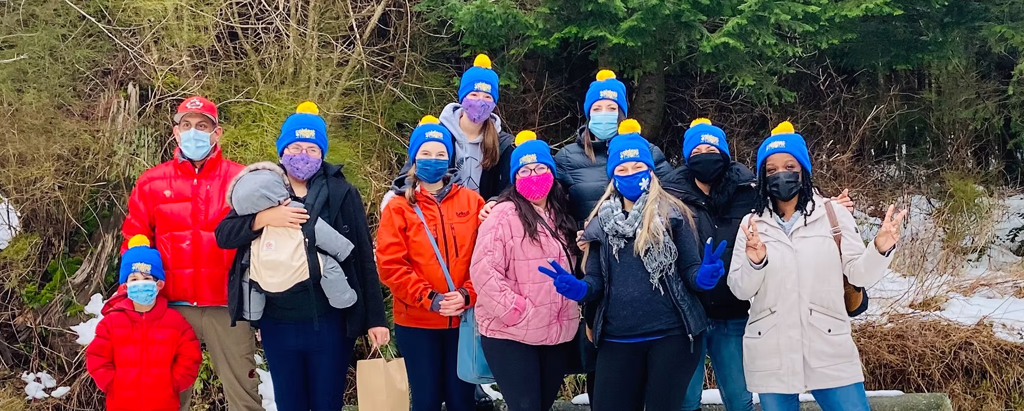 A group of people wearing winter coats, blue hats with yellow pom-poms, and masks stand outside on a snowy ground, posing and smiling for the photo with trees in the background.